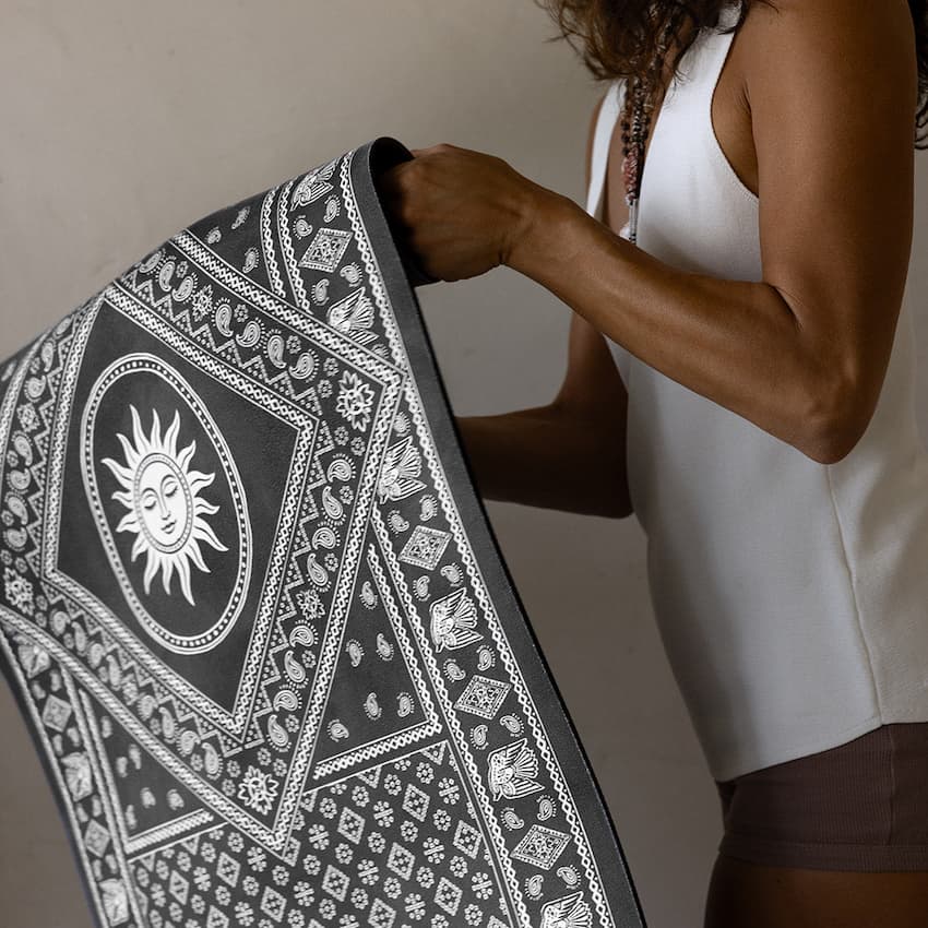 A person holds up a large black and white patterned YOGA MAT  featuring a prominent sun face motif and geometric tribal-style borders.